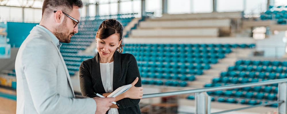 Two Sports Studies students stand in the middle of an empty gymnasium reviewing a playbook together. 